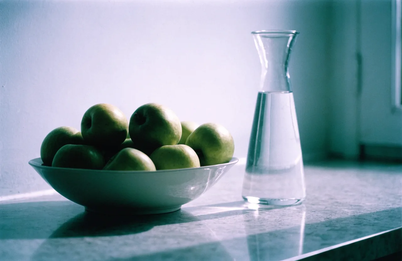 A pale ceramic bowl of green apples and a glass carafe of water on a gray stone counter in soft light.