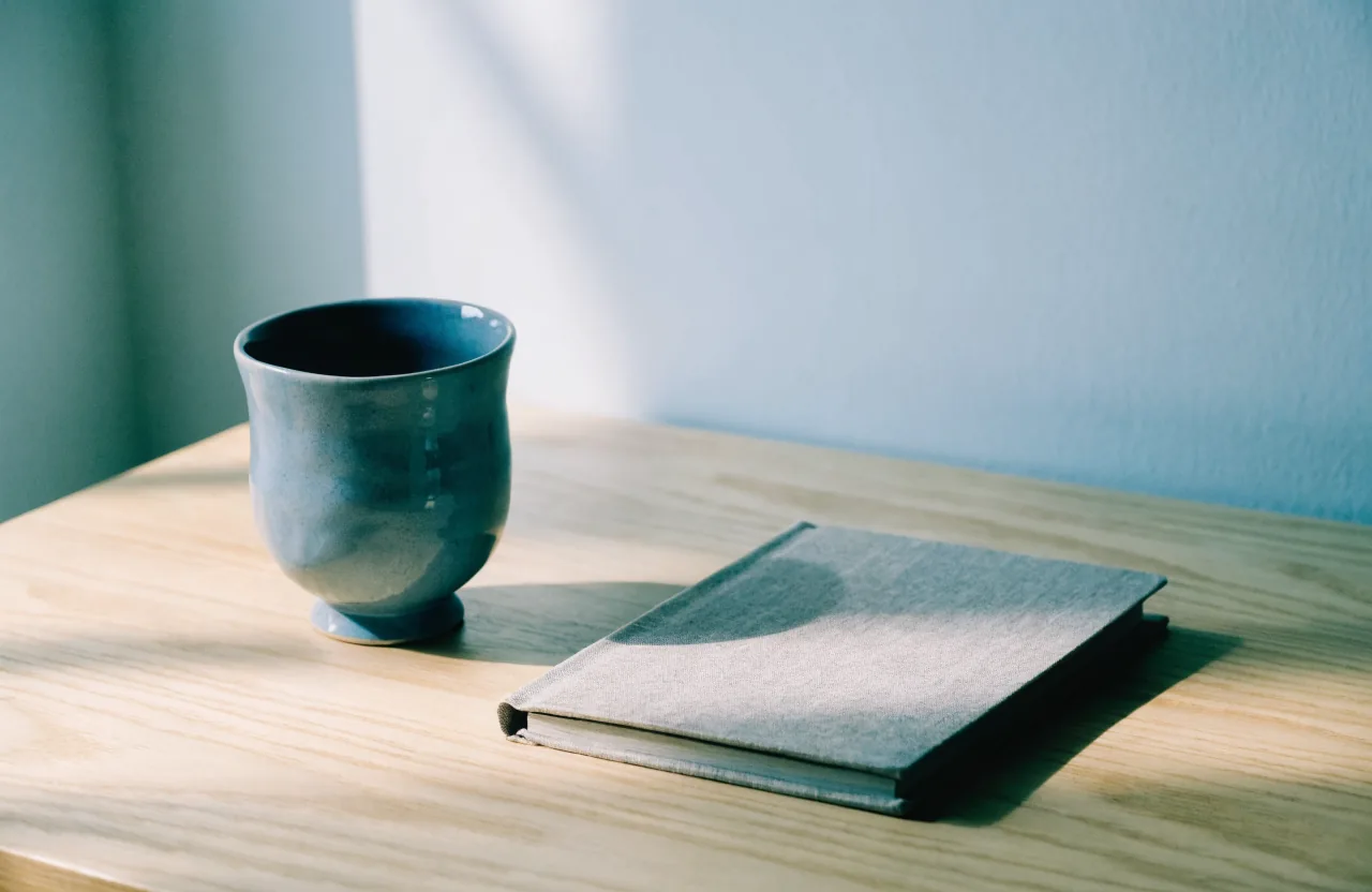 A slate blue ceramic cup beside a closed gray linen-bound notebook on a pale ash desk in cool window light.